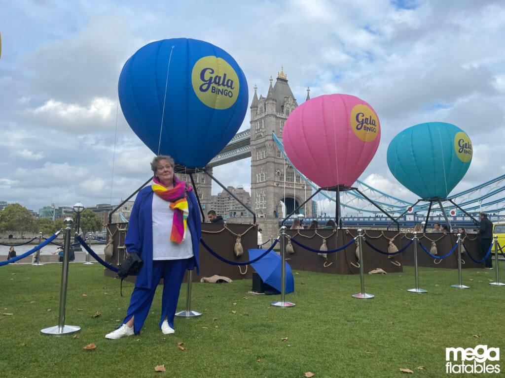 Three colourful branded inflatable hot air balloons with “Gala Bingo” logos displayed outdoors on a grassy area near Tower Bridge, with a person posing in front.